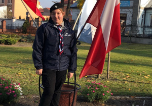 Dziewczynka w mundurze harcerskim stoi na chodniku, a za nią znajdują się flagi Polski.